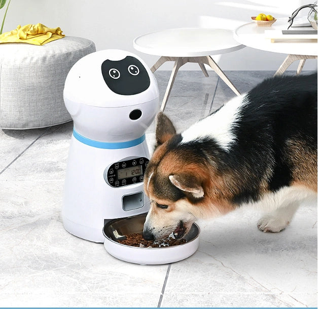 Dog eating from a bowl next to a white electronic pet feeder on a tiled floor.